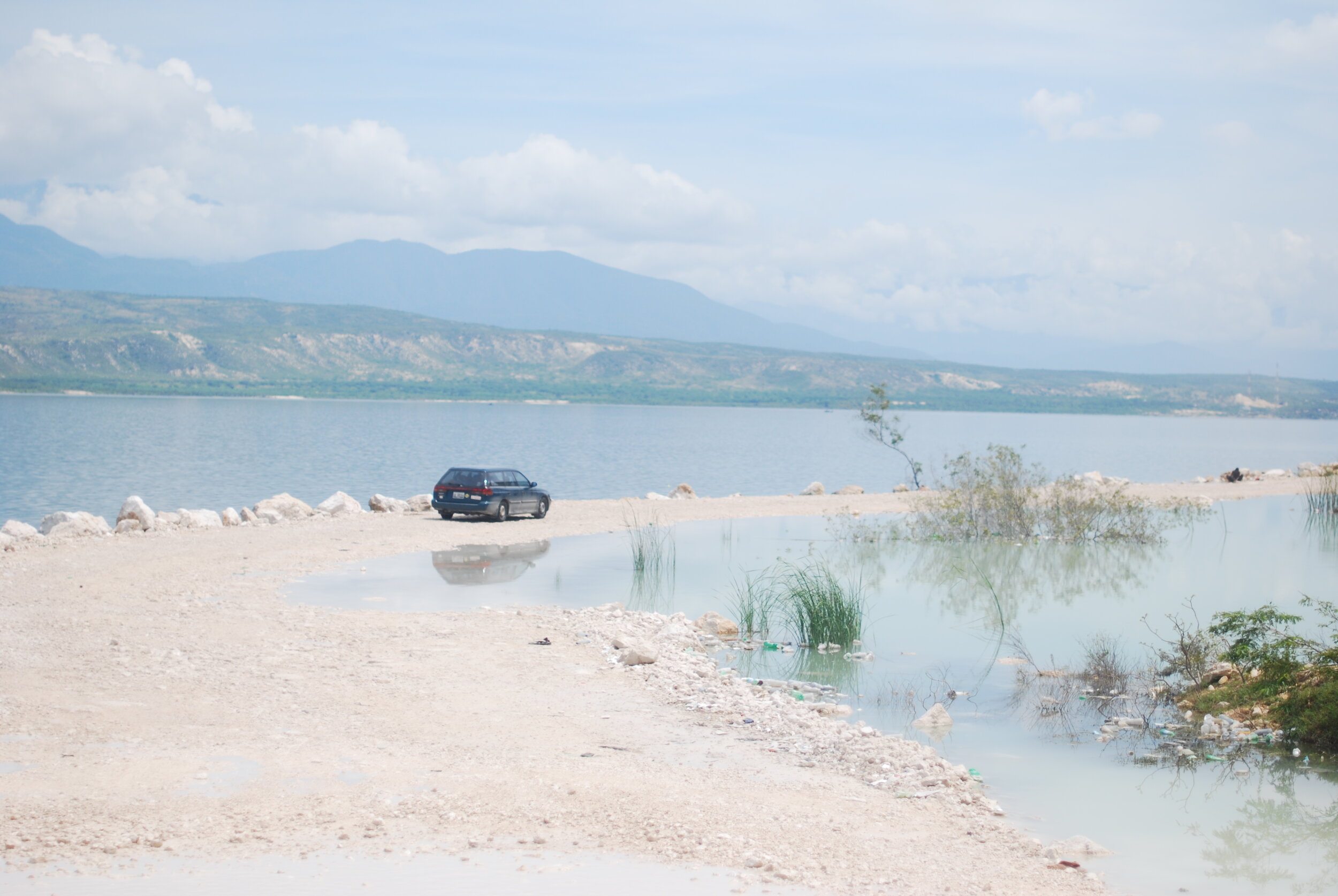 Siri B.L for Wikimedia Commons. Image is of a blue vehicle on the main road from Jimaní, Dominican Republic to Port-au-Prince, Haiti. The pathway is partially flooded by the Lake Azuei.