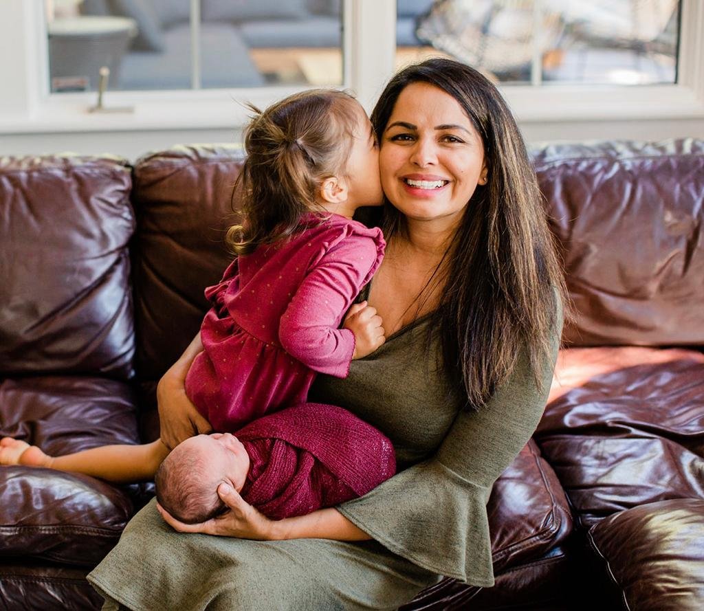 Mahvish sitting on the couch holding her toddler daughter and an infant in her lap. Her toddler is kissing her Mom's cheek while Mom looks to the camera.