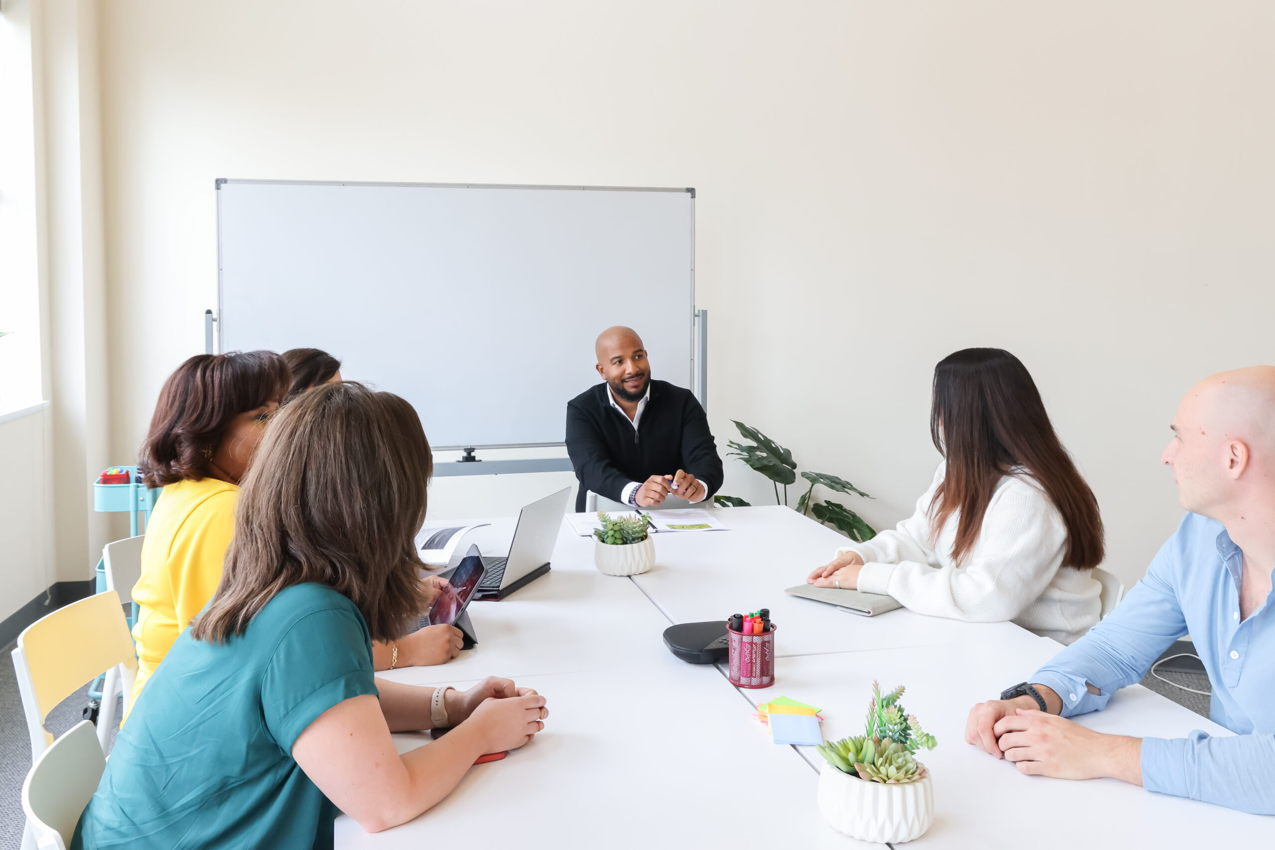 6 people sitting around a desk, in a professional setting. Everyone is looking at the speaker who is sitting in front of a white board,